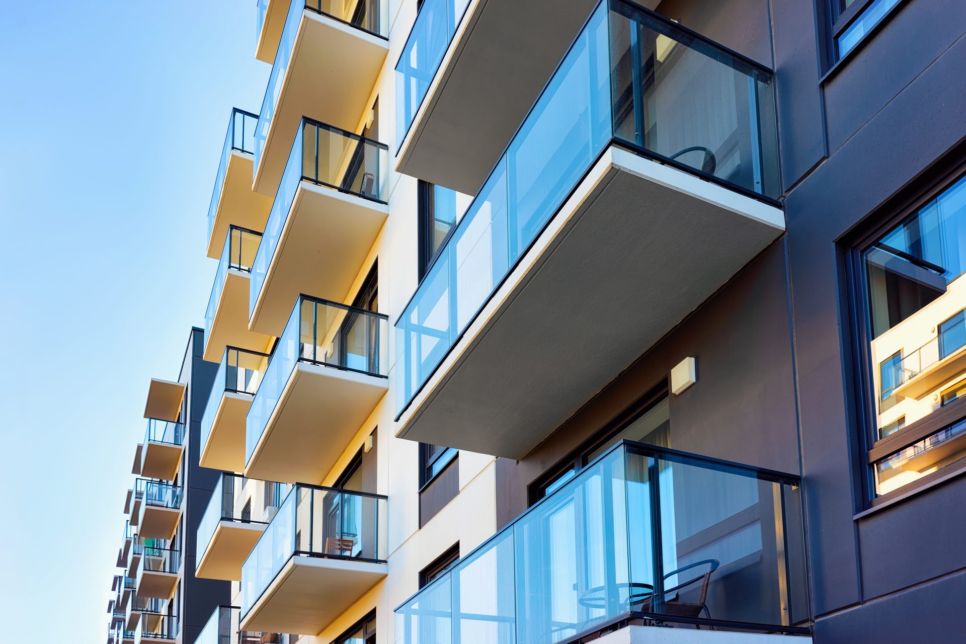 Looking up at a modern apartment block with glass balustrades on every balcony, dark cladding and pale soffits against a blue sky