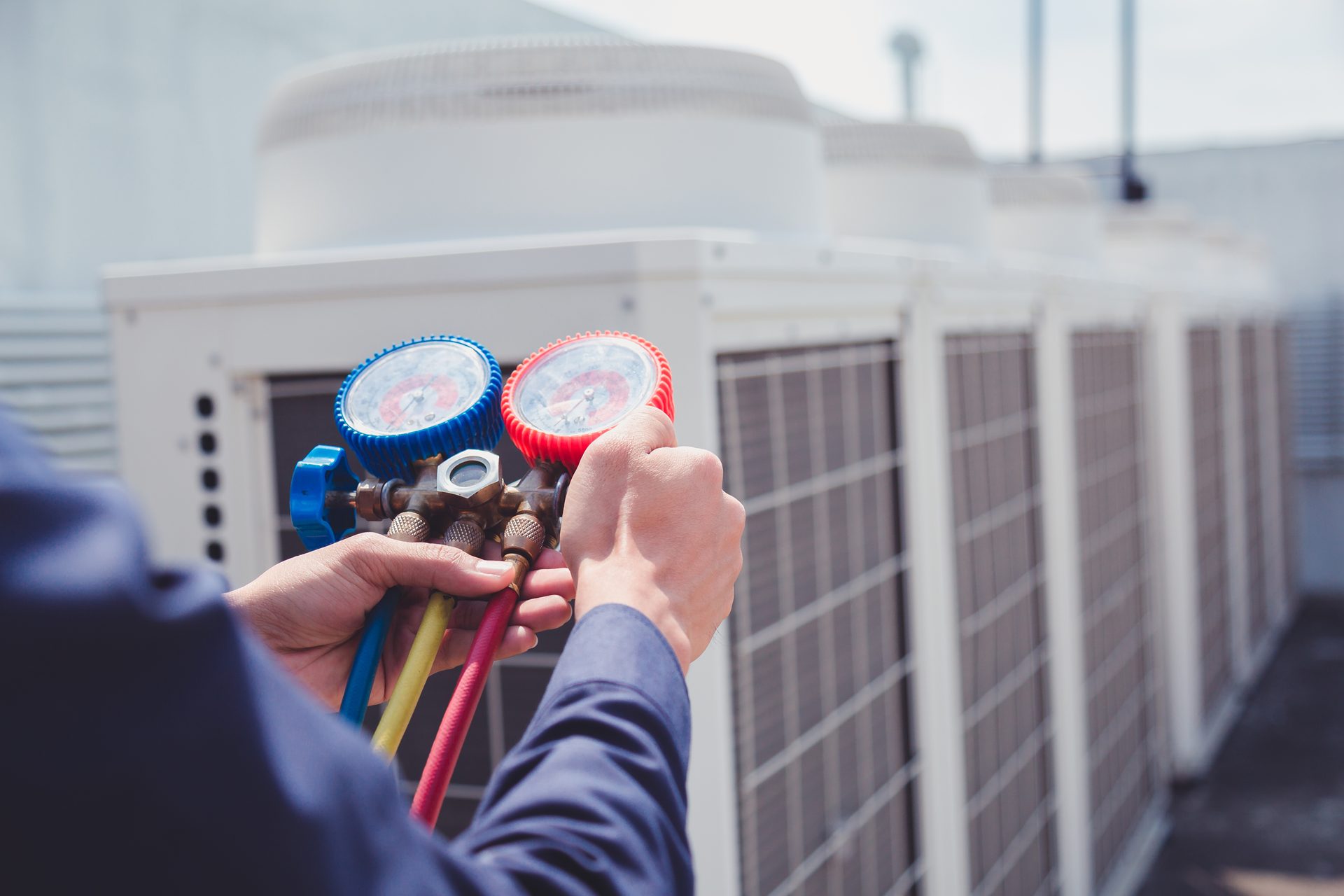 Hands holding a pair of refrigerant pressure gauges beside a row of large outdoor condenser units on a roof