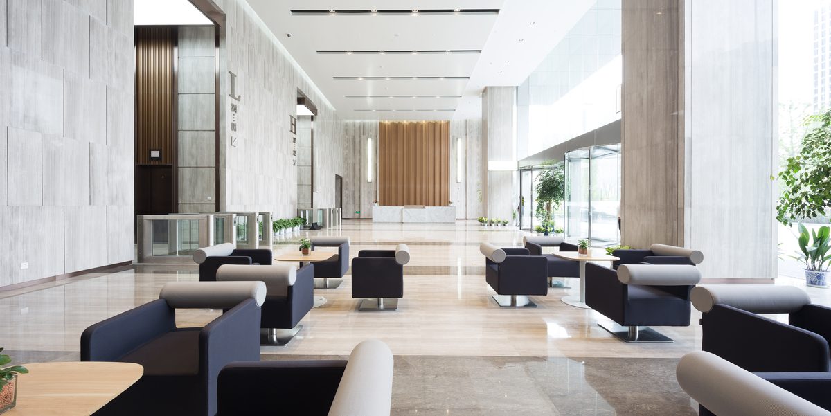 A long building lobby with grey lounge chairs and sofas, pale stone walls, a timber feature wall and tall windows