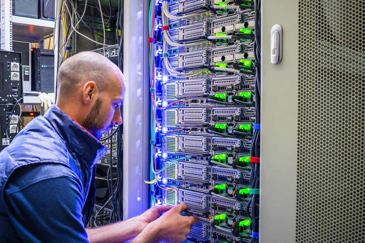 A technician working on the cabling of an open server rack, lit by rows of blue and green equipment lights